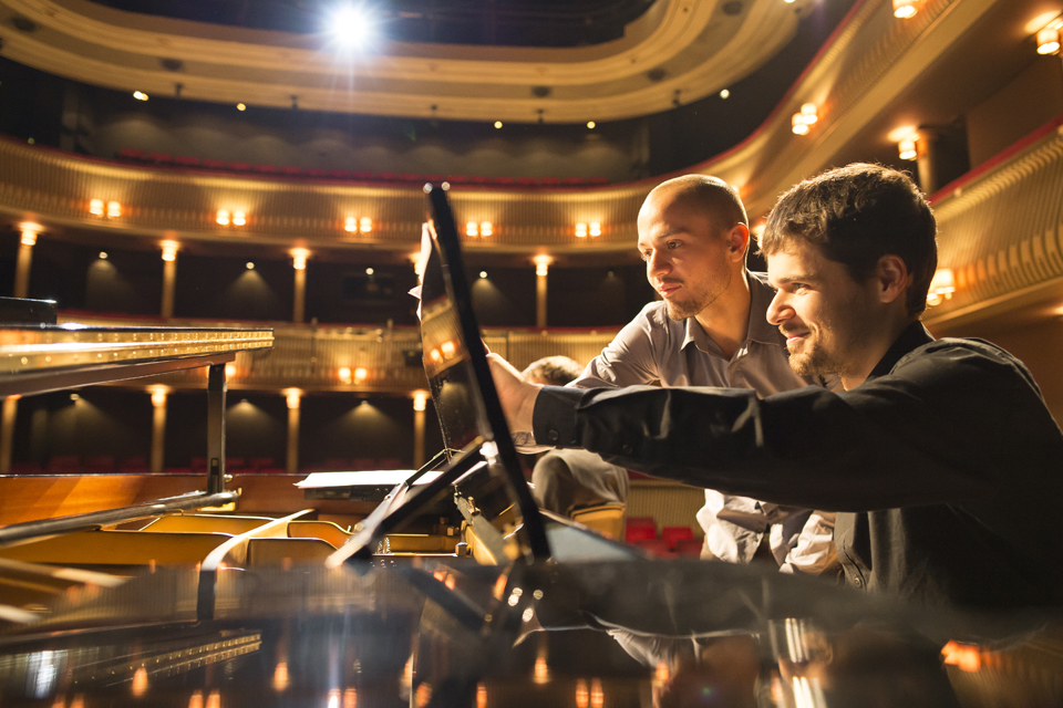 A male student, standing next to a sitting male student, pointing at a music sheet, placed on a piano, with a well-lit theatre in the background.